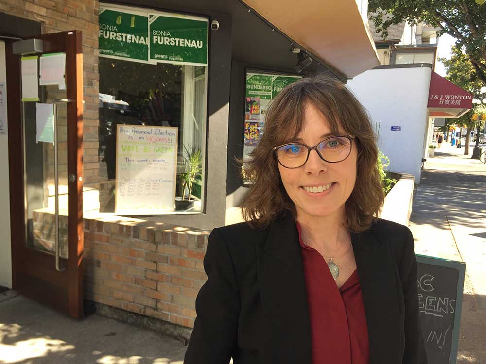 A 50-ish light-skinned woman with shoulder-length brown hair smiles at the camera. She wears glasses and a black suit jacket and burgundy shirt.