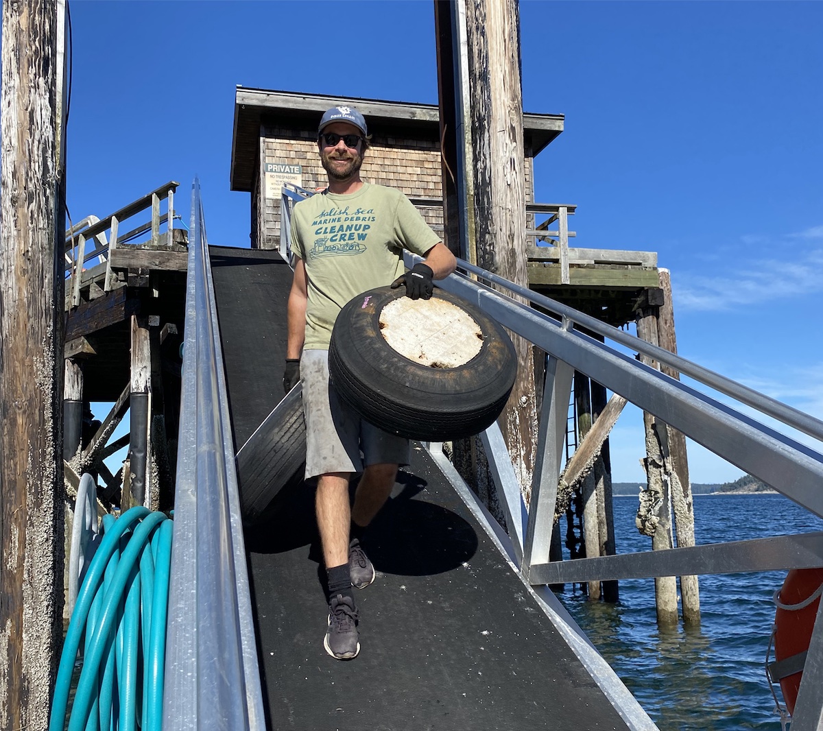 A person in a blue ball cap and green T-shirt carries a tire filled with white Styrofoam down a black walkway to a dock.