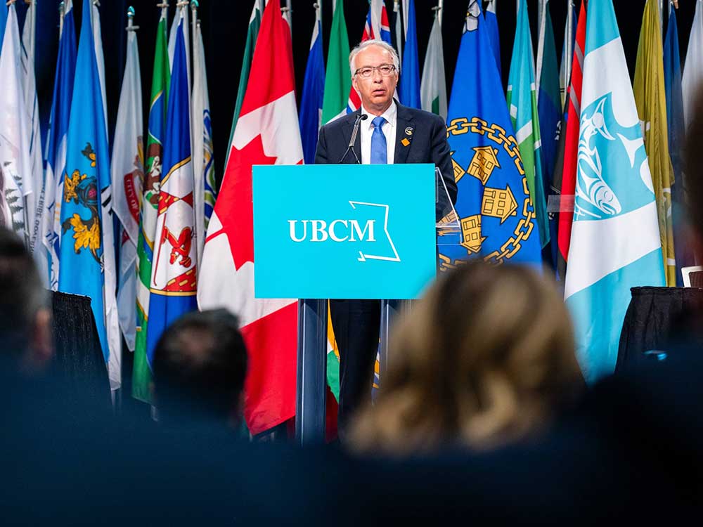 John Rustad is a white man with short grey hair and glasses. He is wearing a navy suit, light blue tie and white button-down shirt. He stands at a podium with a turquoise UBCM sign on it. Behind him is a colourful row of flags, and a crowd of audience members is in the darkened foreground.