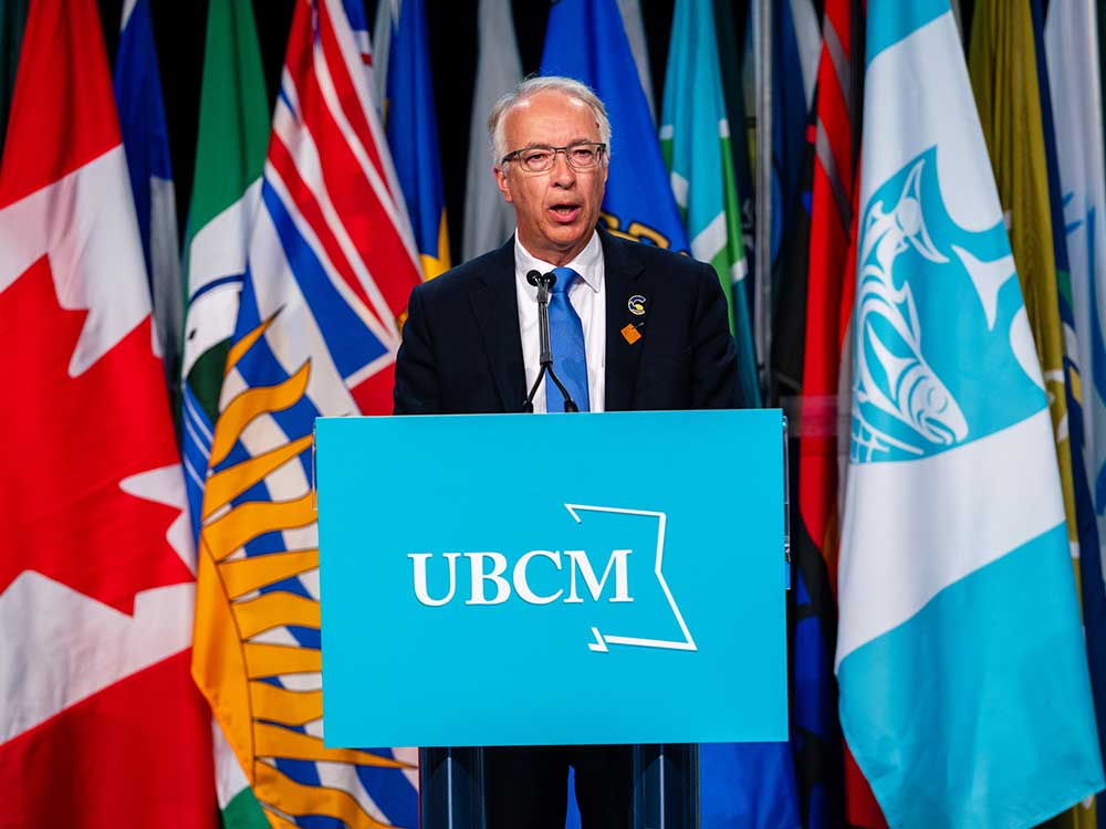 A 60-ish light-skinned man in a suit stands at a podium reading 'UBCM.' He is surrounded by flags.