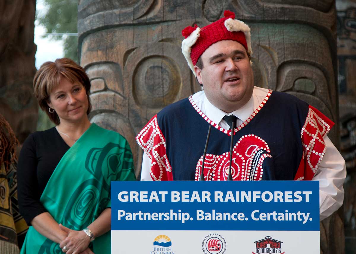 A man wearing traditional Indigenous clothing stands at a podium that says 'Great Bear Rainforest' and speaks into a microphone. On the left, a woman with short, dark hair wearing a green scarf with an Indigenous design looks on.
