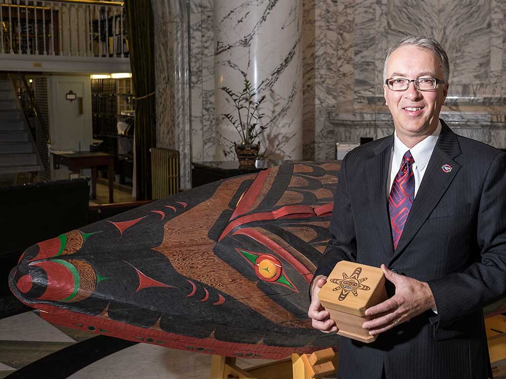 A man with short, grey hair wearing a dark suit and glasses holds a wooden box with an Indigenous design on the lid. He stands in a room with marbled walls and there is a large Indigenous-style carving behind him.