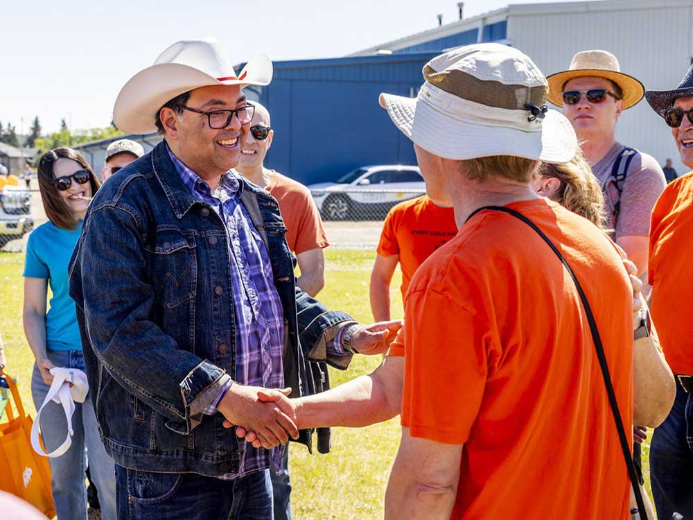 A smiling 50-ish man with medium-toned skin, wearing a white cowboy hat and denim jacket, talks with people under sunny skies.