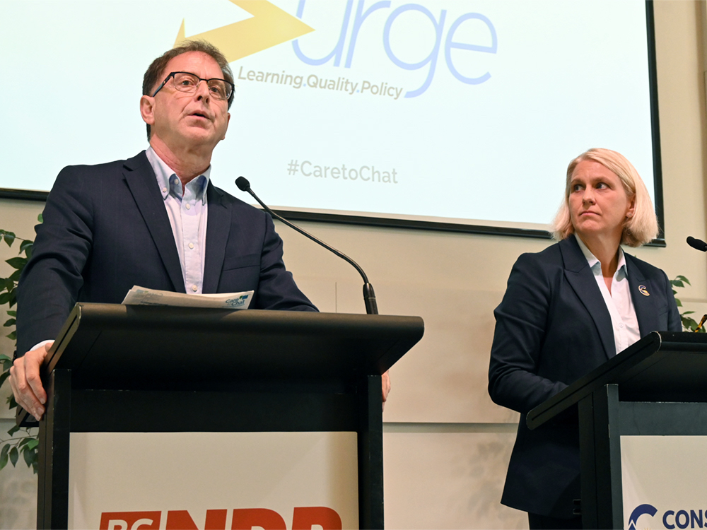 On the left, Adrian Dix, a middle-aged man with light skin tone and brown hair, stands at a podium. On the right, Elenore Sturko, a middle-aged woman with light skin tone and a blond bob, stands at a podium.