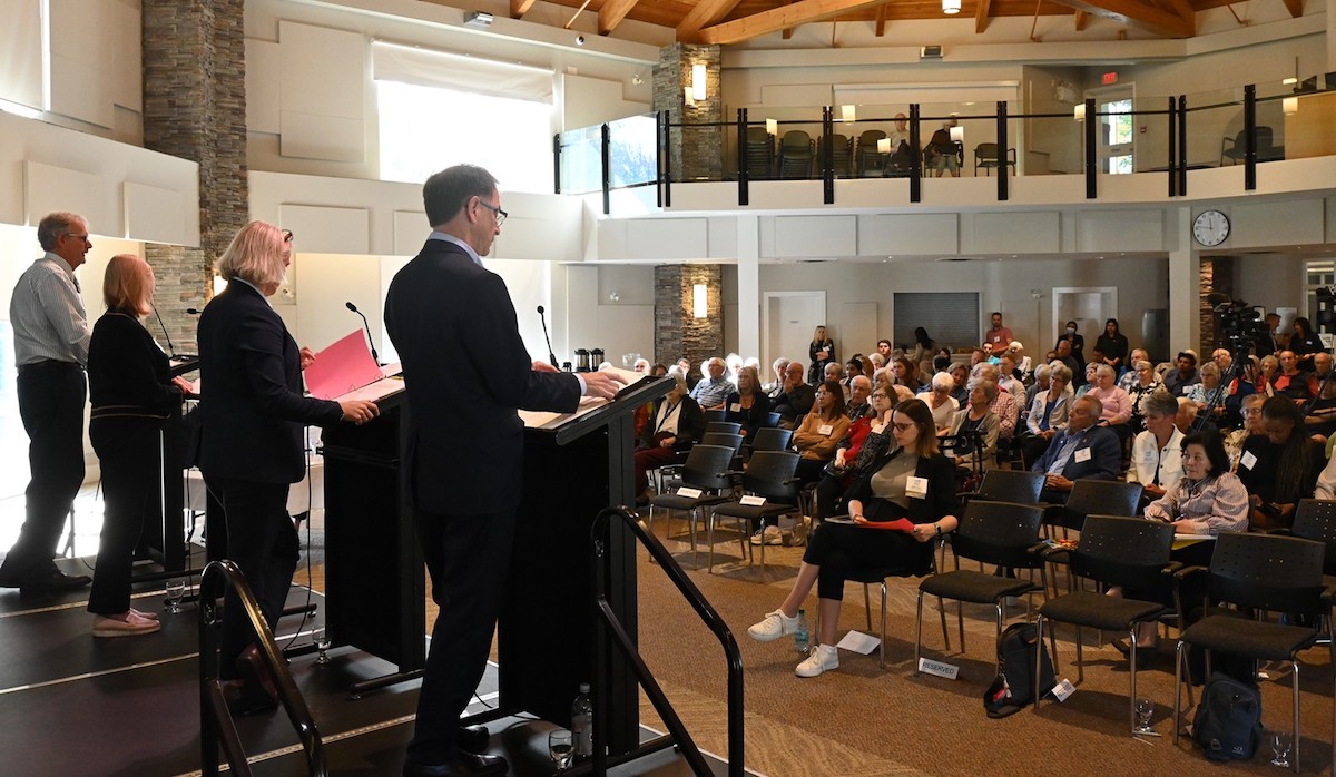Four people stand at podiums on a stage in front of an audience in a small auditorium.