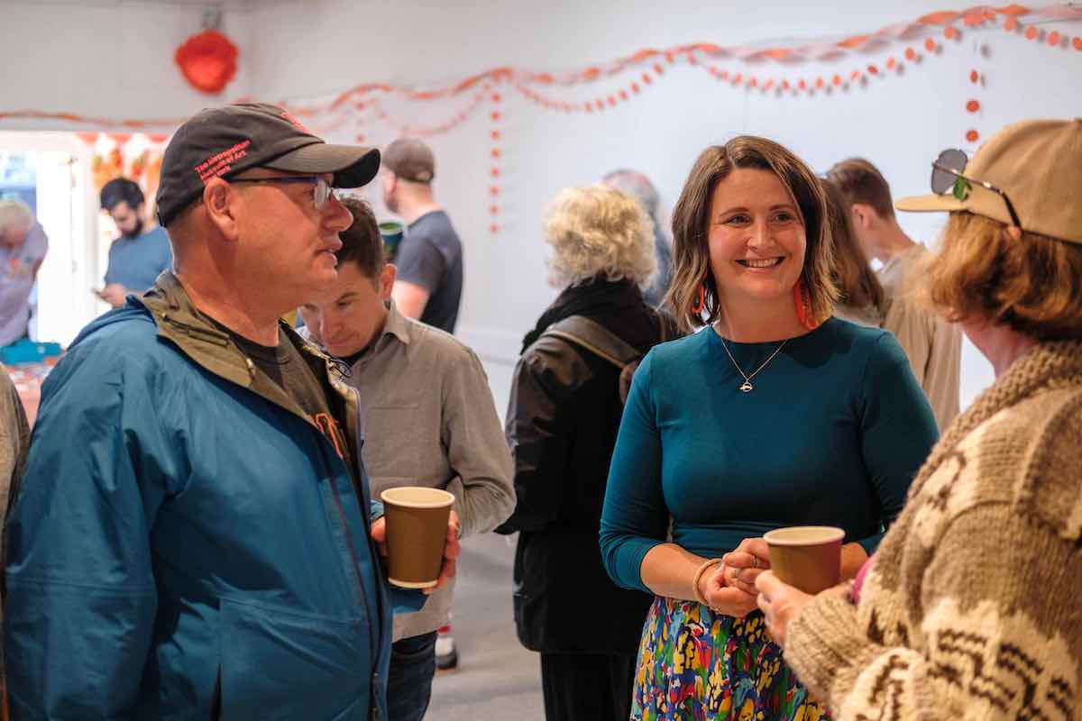 Grace Lore wears a teal top and colourful skirt. She speaks with a person in a thick wool sweater and ball cap to the right of the frame. A man in a ball cap, windbreaker and glasses stands to the left of Lore. Both people are holding coffee cups and they are in an indoor space decorated with orange banners.