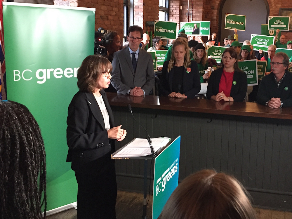 BC Green Leader Sonia Furstenau, a 54-year-old light-skinned woman, stands at a podium with supporters holding green candidate signs in the background.