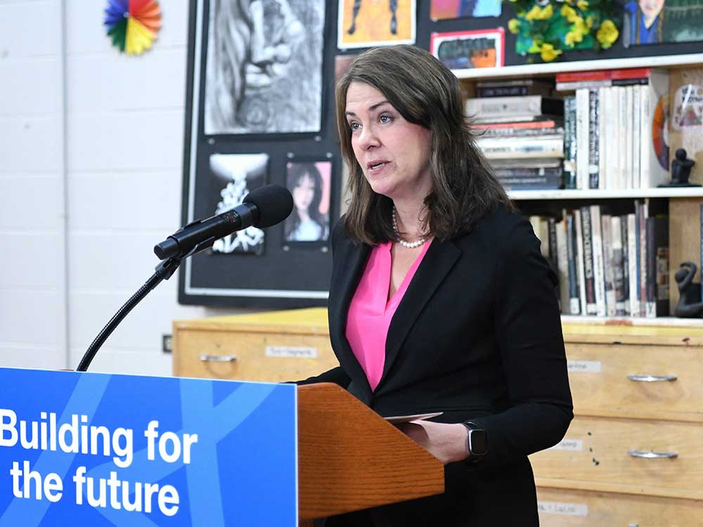 A 50-ish light-skinned woman with shoulder-length brown hair stands at a podium.