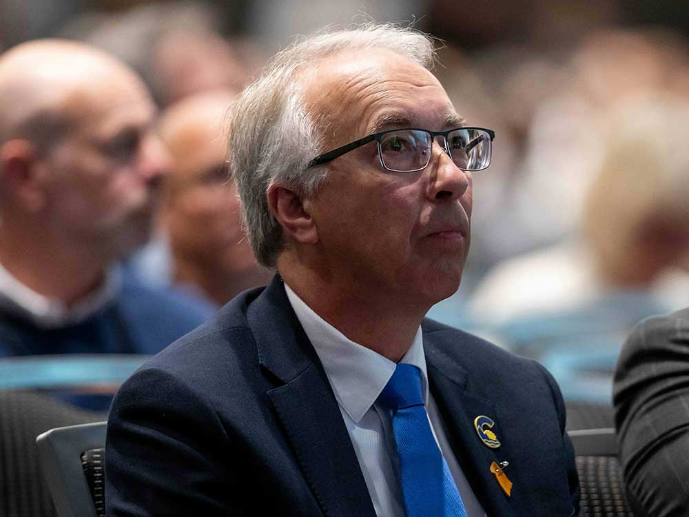 A 60-ish light-skinned man with grey hair wears a blue suit. On the lapel are a Conservative Party of BC badge and a Moose Hide pin.