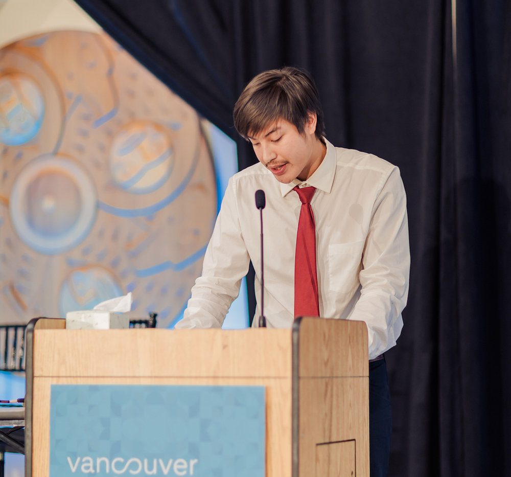 A young man with medium-light skin tone and brown hair speaks at a podium. He is wearing a white collared shirt and red tie.