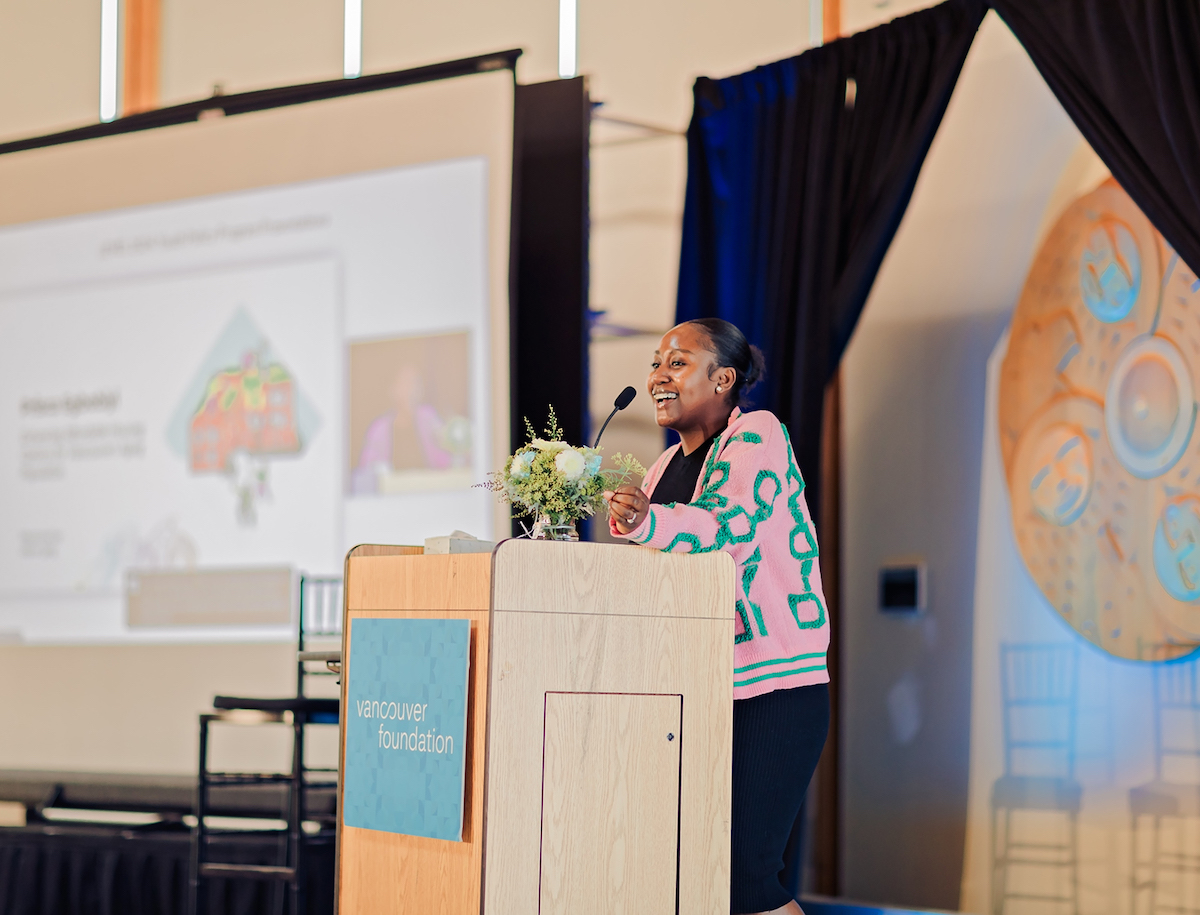 A young woman with medium-dark skin tone stands at a podium addressing an audience. She is wearing a pink cardigan with raised turquoise detailing.