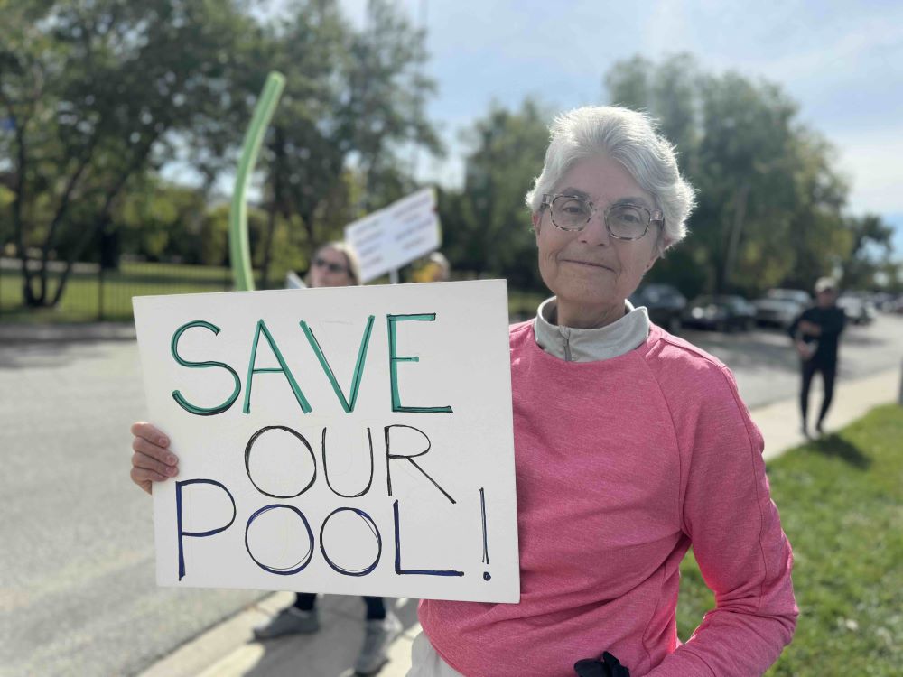 Betty Leinweber has short grey hair and glasses. She is wearing a pink top and holding a handmade sign that reads &ldquo;Save our pool.&rdquo;
