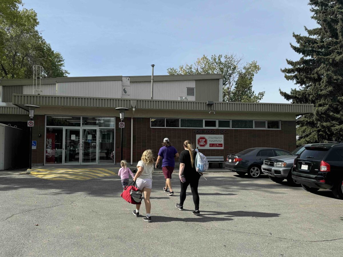 A family of four walks with their backs to the camera through a parking lot towards the front entrance of the Inglewood Aquatic Centre. They are holding bags of swim towels and gear. It&rsquo;s a sunny day.