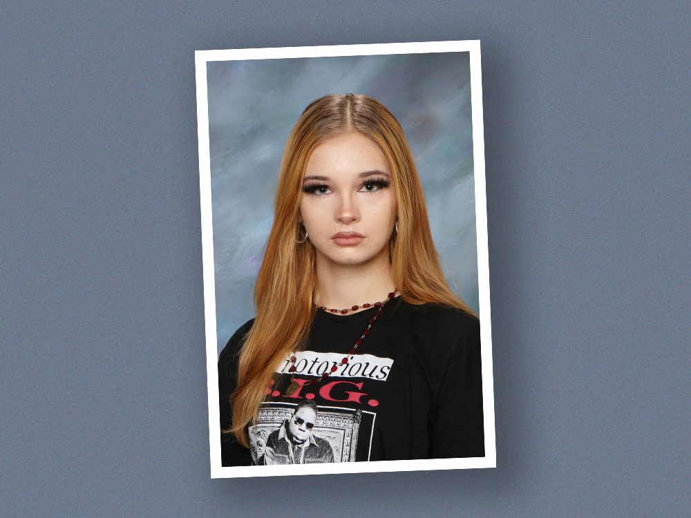 A school photo of a teenage girl with light skin tone and long brown hair. She is wearing a Notorious B.I.G. shirt.