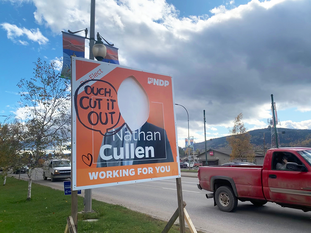 A large orange sign that says 'Re-elect Nathan Cullen' stands by the roadside. There is a hole in the middle of the sign where a person’s face has been cut out. 'Ouch, cut it out' is written in black marker.