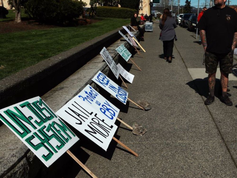 Placards with text reading "Un-just system," "Jail more" and "Keep in custody" sit propped up on a concrete sidewalk and curb. 