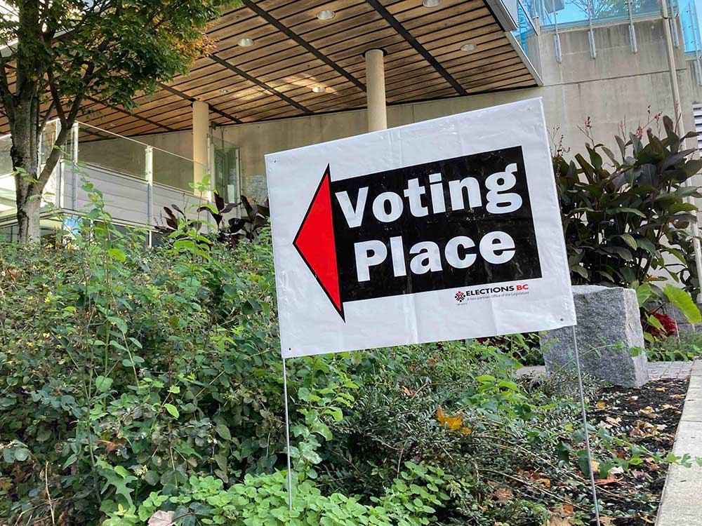 A sign with white sans-serif text over a black rectangle, red arrow and white background reads 'Voting Place' with greenery and a modern glass and concrete building behind it.