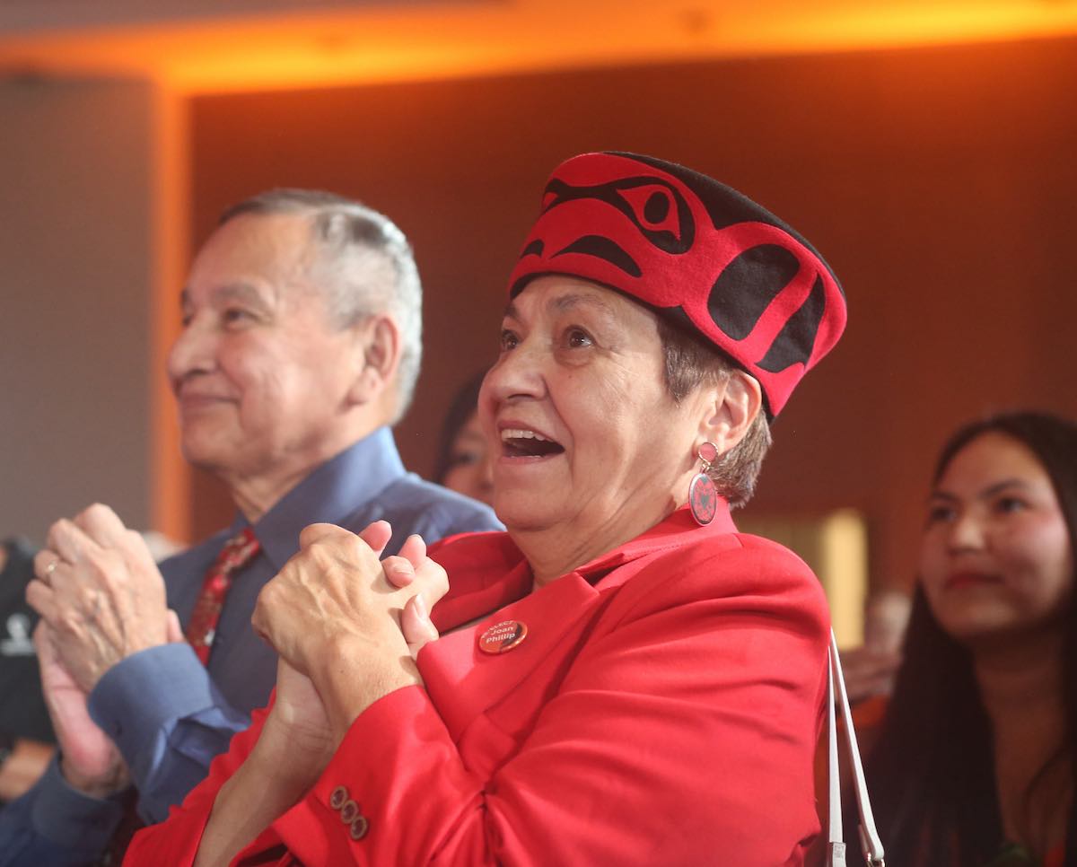Joan Phillip, wearing a black and red hat with Coast Salish design and a red blazer, clasps her hands and looks excited.