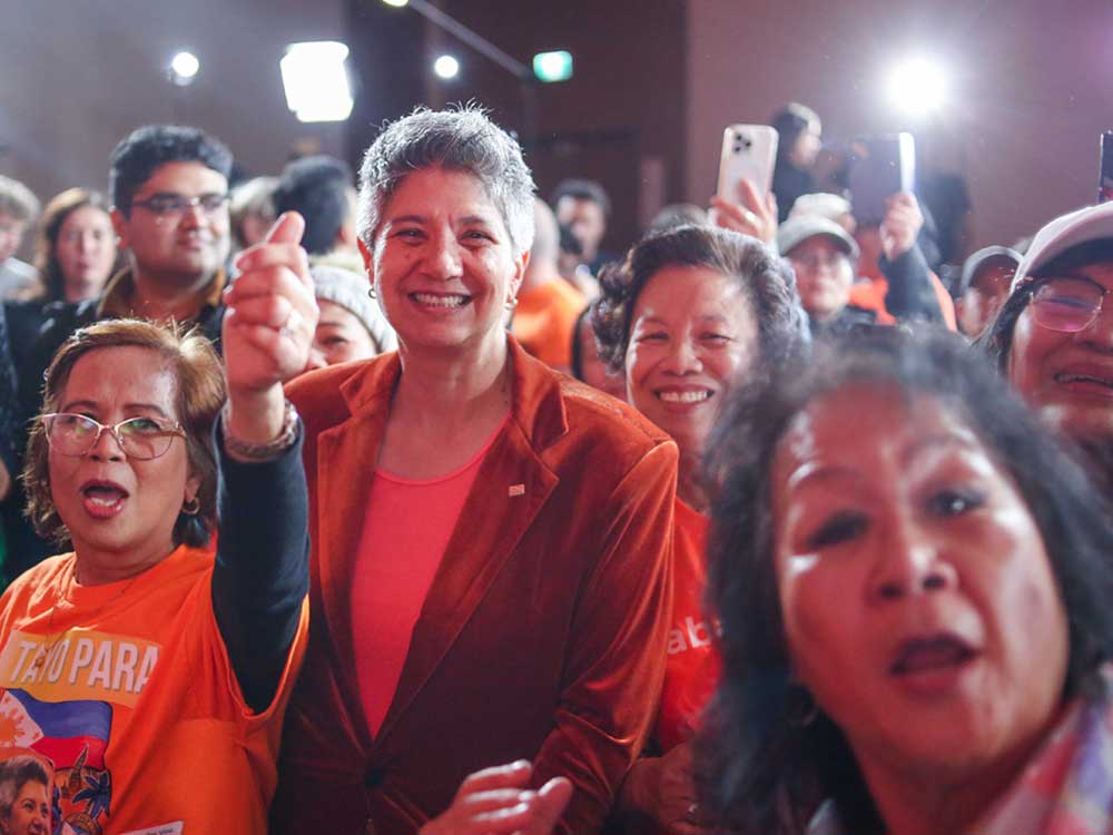 Mable Elmore, a middle-aged woman with medium skin tone and short dark grey hair, wears an orange shirt and an orange velour blazer and smiles. She&rsquo;s surrounded by supporters wearing orange.