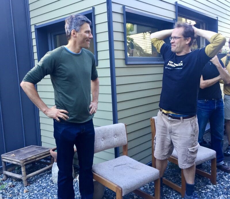 Two men stand in conversation in front of a home.