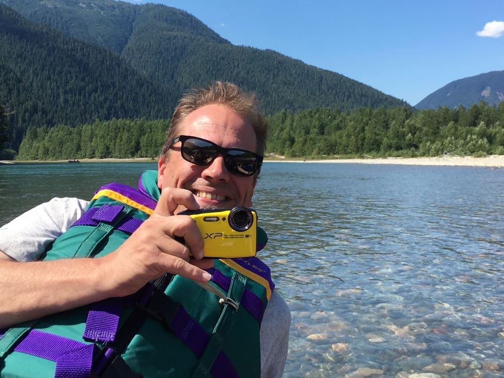 A man with light skin and light brown hair wearing sunglasses and a life jacket takes a selfie with a clear lake and tree-covered mountains behind him.