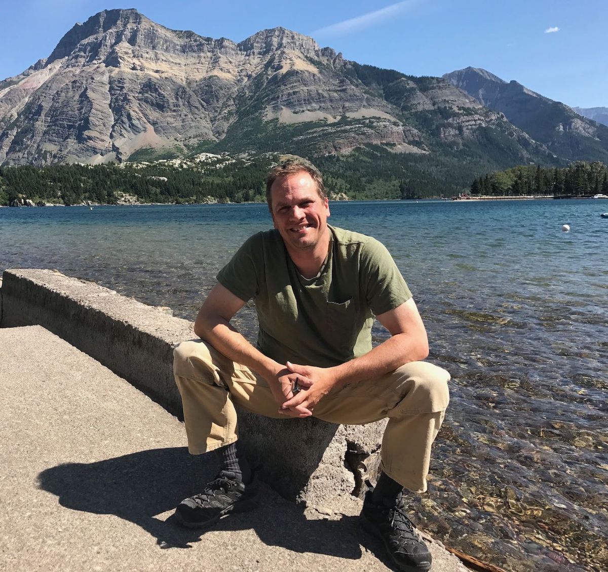 A man with light skin and light brown hair, wearing a green T-shirt, khaki pants and black hiking shoes, sits on a low concrete wall by a lake, with a rocky mountain in the background.
