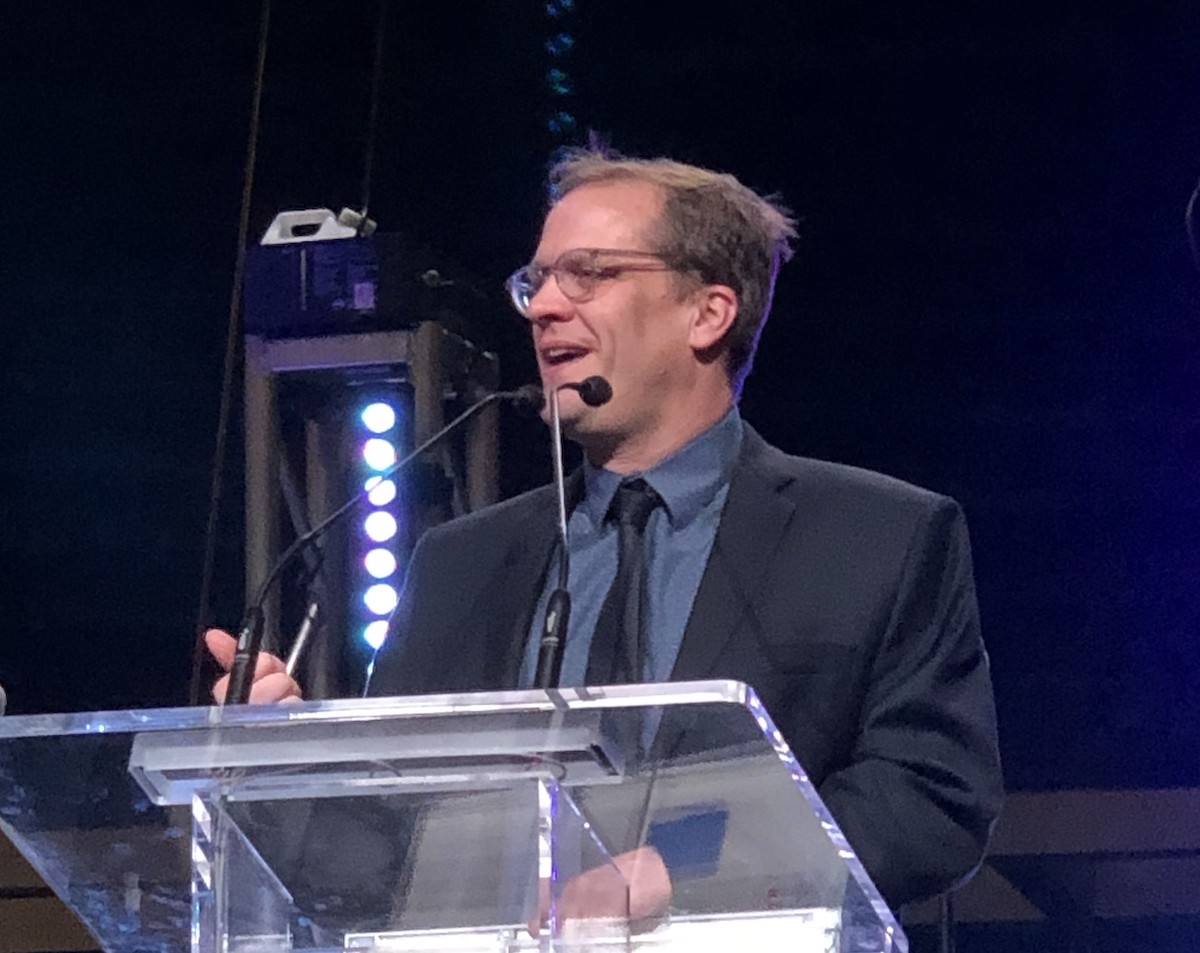 A man with light skin and light brown hair, wearing glasses, a black jacket and tie and a blue shirt, speaks at a podium.