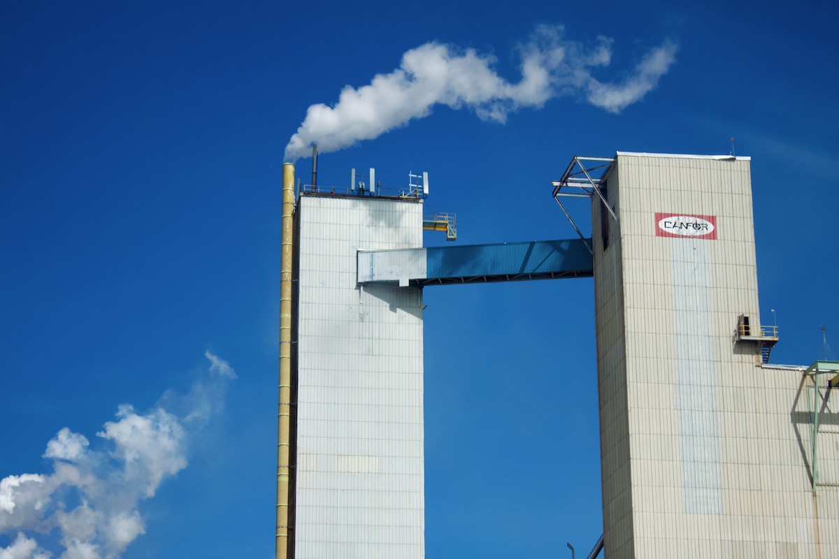 A large, mostly grey pulp mill. It has a large 'Canfor' sign and two smokestacks painted red and white.