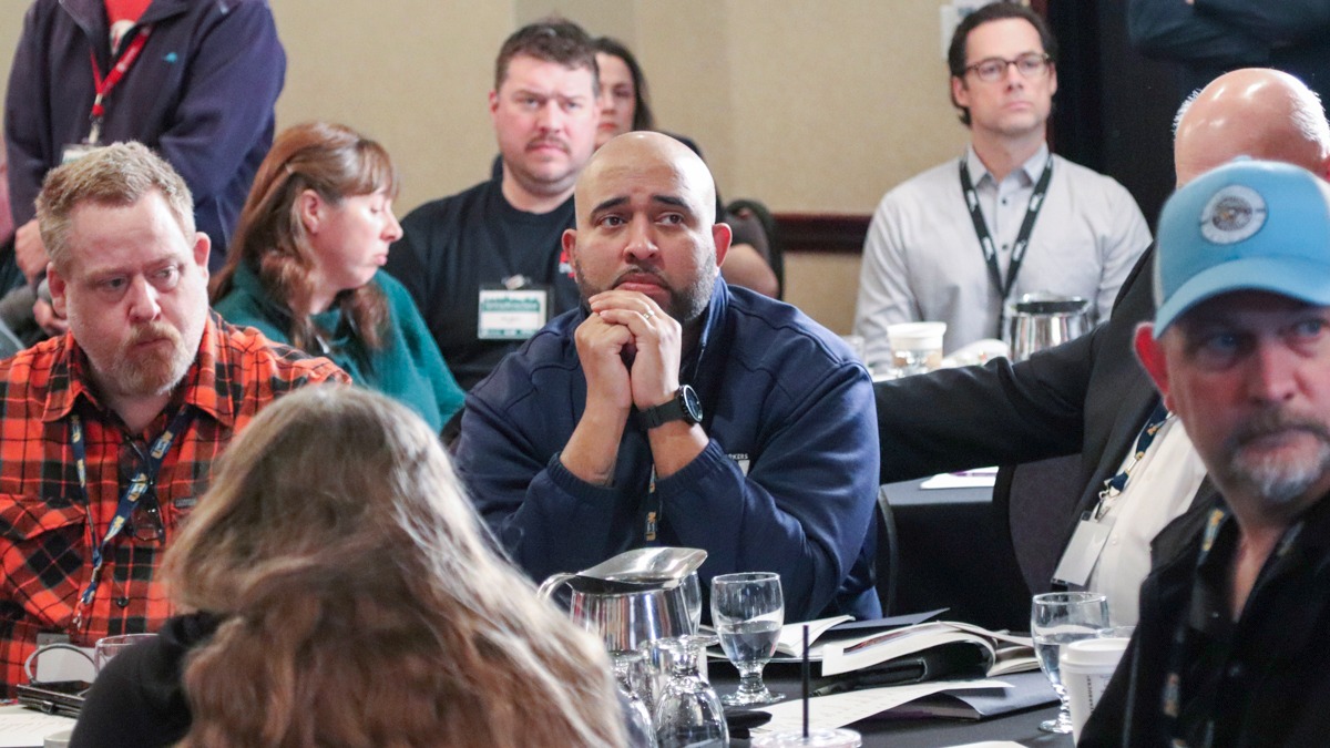 Groups of men, with some women interspersed, wear lanyards and sit at round tables.