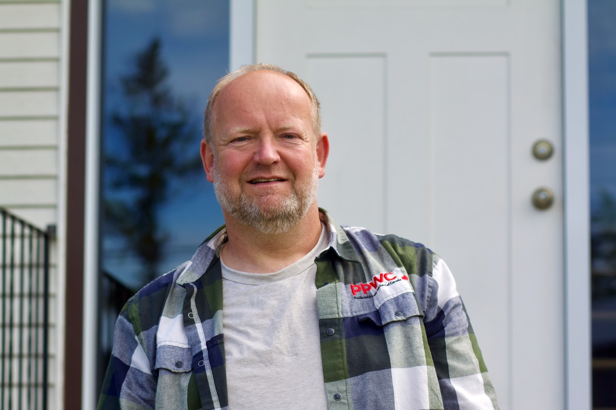 A balding man with light skin tone stands outside a house, wearing a plaid shirt embroidered with 'PPWC.'
