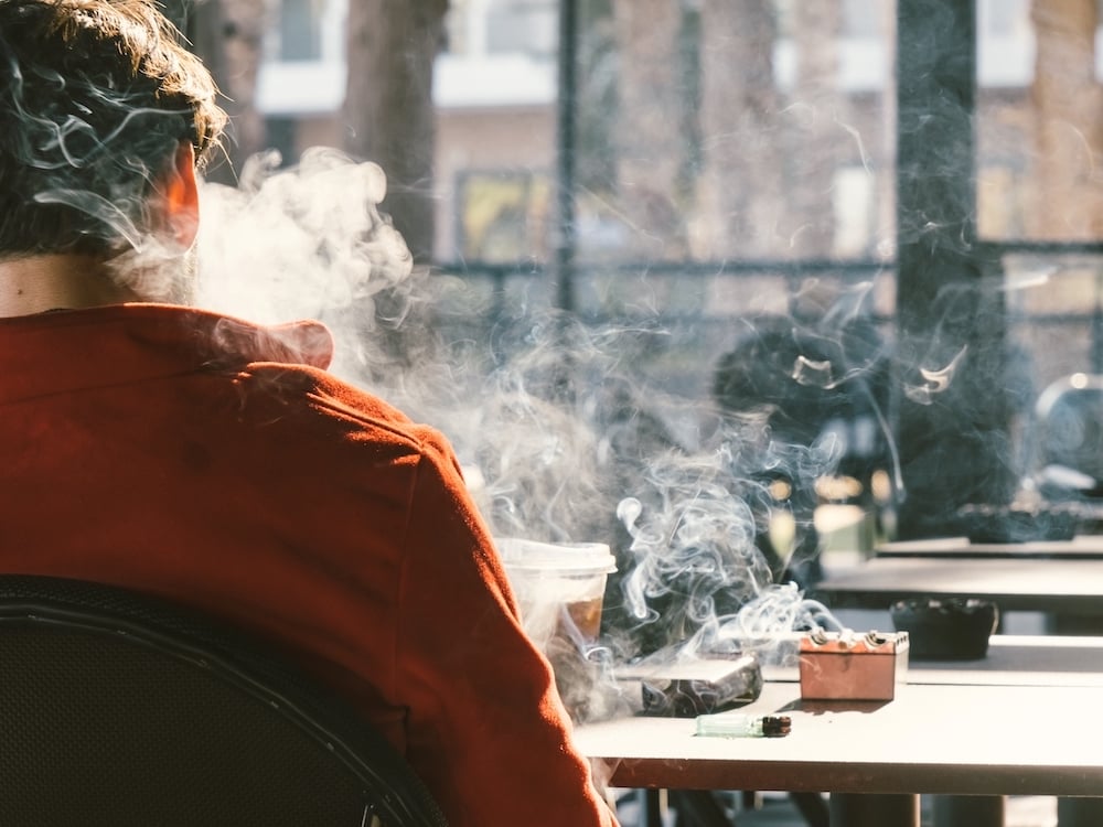 A man sits at a table outdoors, his back to the camera. He is smoking. There’s a cloud of smoke and an ashtray.