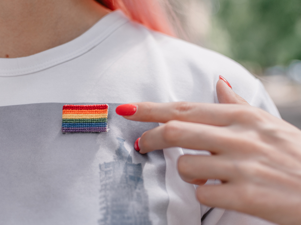 A hand with light skin colour and painted red nails points to a beaded rainbow Pride flag pinned to their white and grey T-shirt.