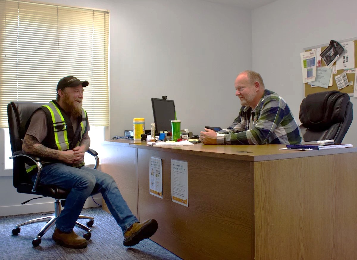 On the left, Jonathan Blacker, a man with light skin tone, a long orange beard and tattoos, wearing a safety vest, sits across a desk from Greg Petersen, a balding man with light skin tone who is wearing a plaid shirt.