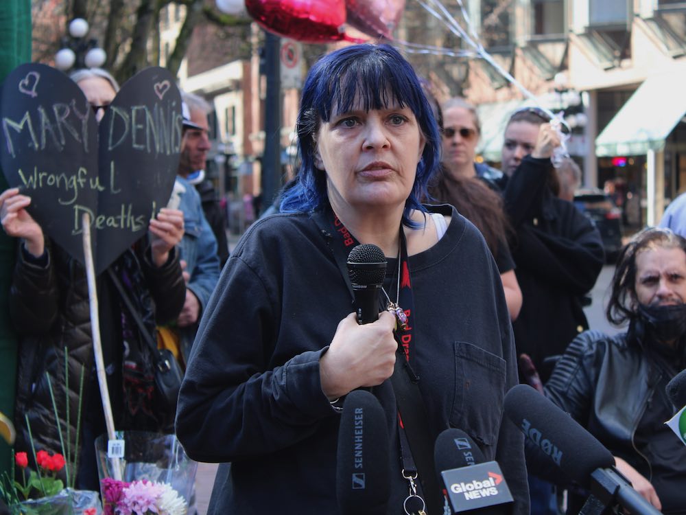A light-skinned person with purple hair, wearing a black shirt and holding a microphone, speaks to media.