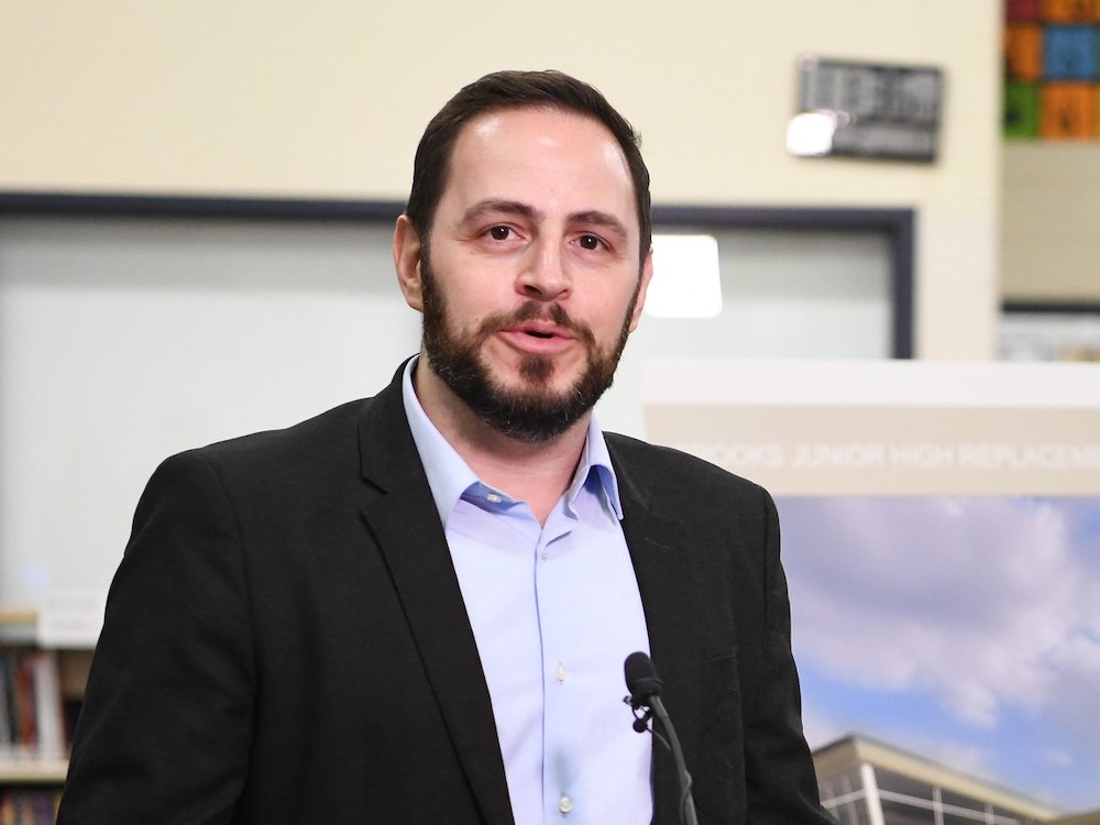 A man with light skin, short dark hair and beard, wearing a dark suit jacket over a light blue collared shirt, looks into the camera while speaking into a microphone.