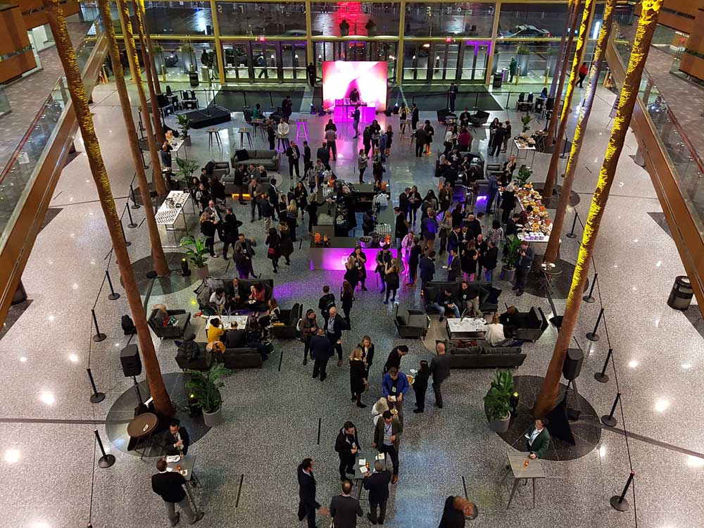 A photo taken from an upper mezzanine looks down upon a crowd mingling over drinks in an atrium at the 2018 CityLab conference. People in business wear stand in small groups at high-top tables or are seated together on black sofas. At the front of the room, a DJ is stationed at a set of turntables against a brightly lit magenta screen.