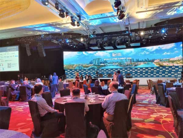 People are seated around circular tables at a conference. On the wide screen at the front of the room is a panoramic photo of an urban landscape on the water. White sans-serif text reads 'World Cities Summit 2022.'