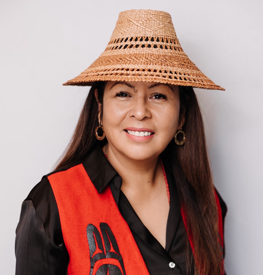 A 40-ish Indigenous woman smiles at the camera. She wears a woven cedar bark hat, black shirt and red vest.