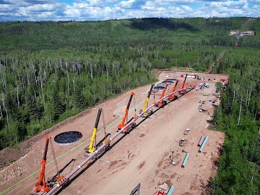 An aerial photo shows forests, a large cleared patch and a row of eight large cranes lifting a long section of pipeline.