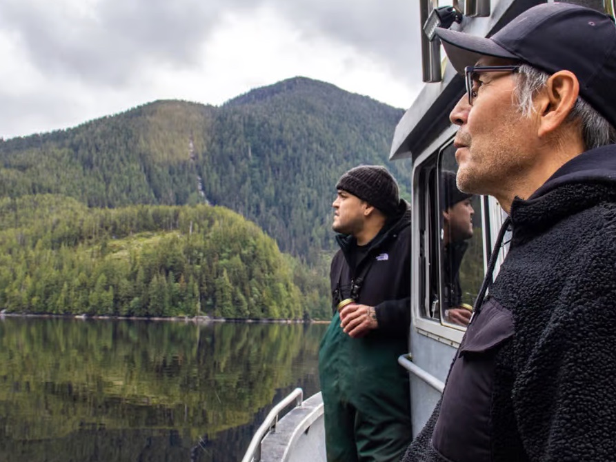 Two Indigenous men stand on an aluminum work boat looking into the distance. They are on smooth water with forested hills behind them.