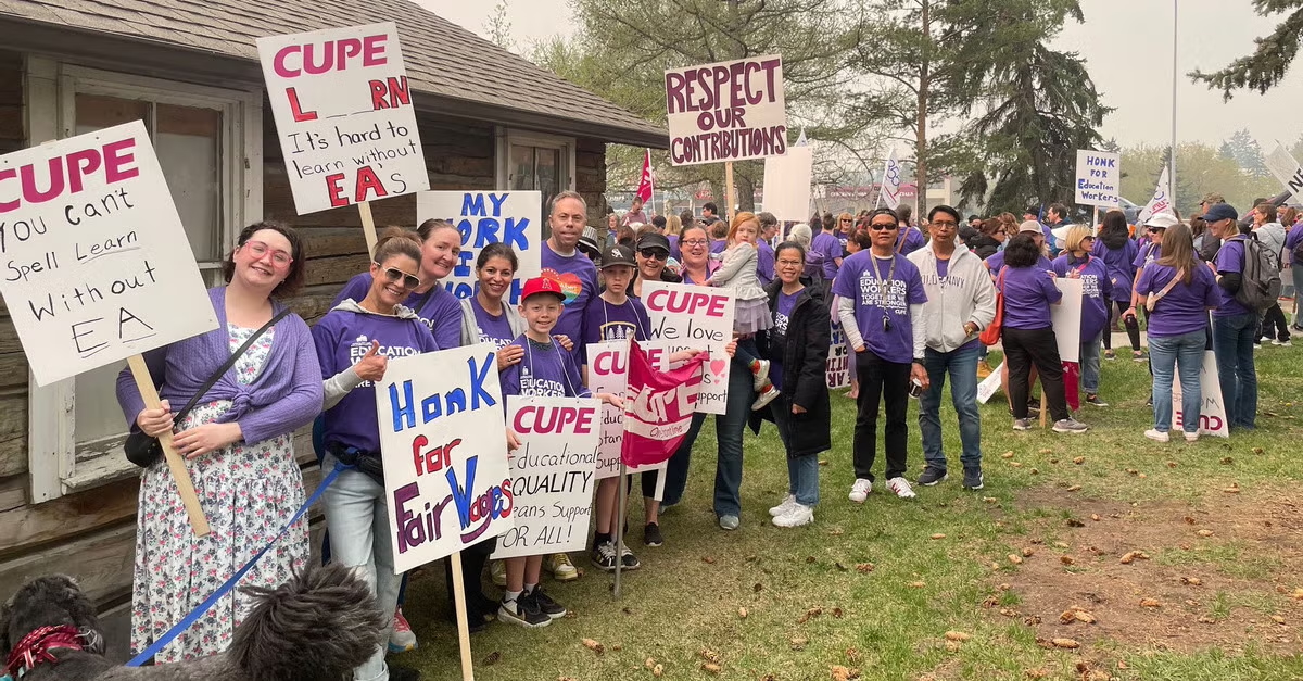 A group of people, some children, stand outside a building. Most are wearing purple and holding signs that call for support for education assistants.