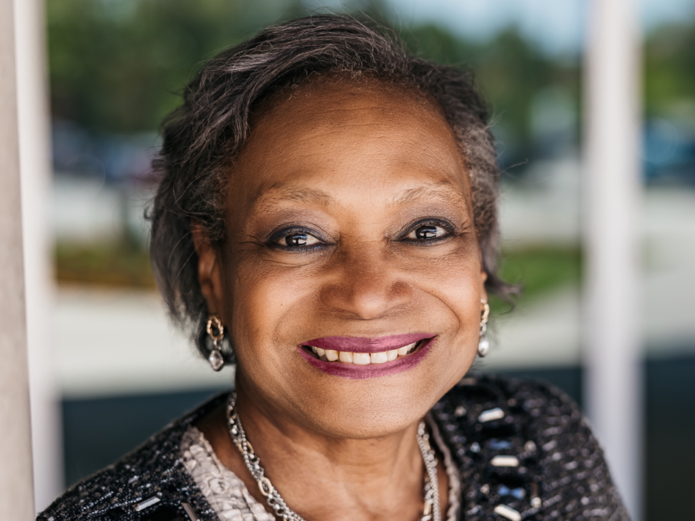 A woman with medium-dark skin tone and short, salt-and-pepper hair smiles at the camera. She is wearing a black cardigan, several silver necklaces and gold earrings.
