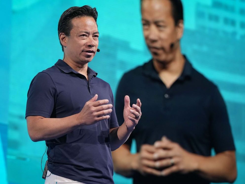 A 54-year-old man of Chinese descent speaks in front of a large screen that shows his image. He wears a blue short-sleeved shirt.