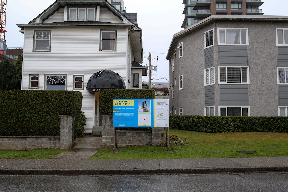 Two three-storey walk-up apartment buildings stand side by side; the one on the left has a white stucco exterior and has features that resemble a home; the one on the right is grey and more contemporary. Between the two buildings is a yellow and blue sign announcing a rezoning application.