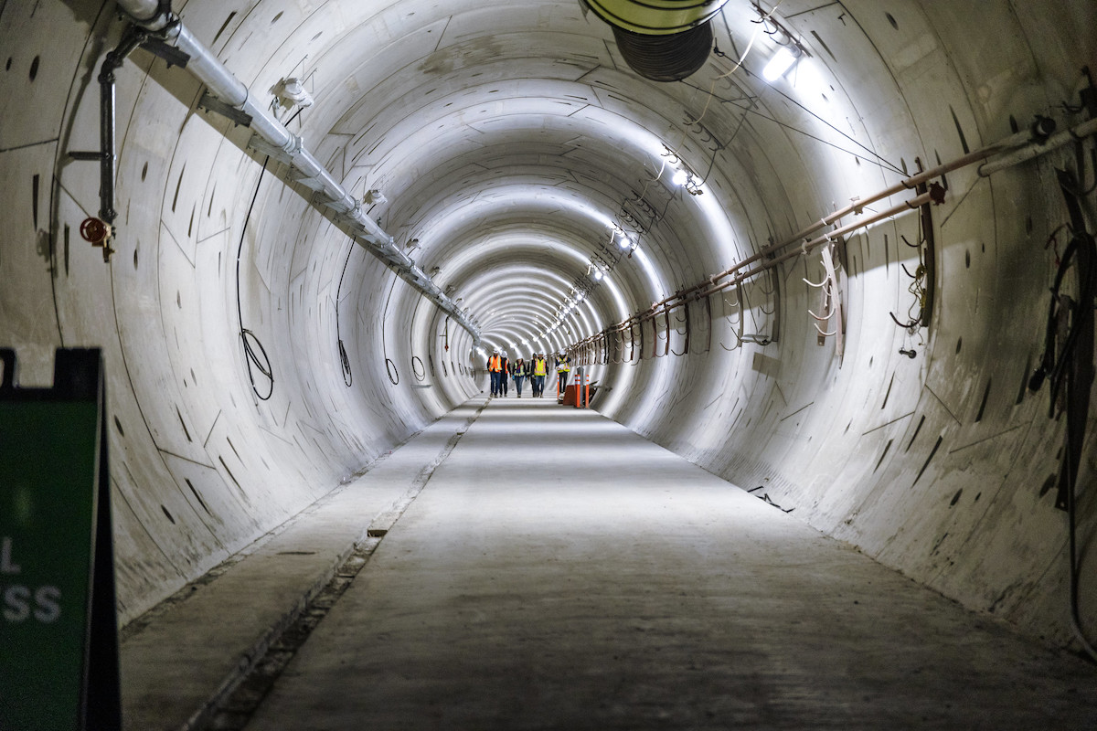 A group of people in high-visibility vests walks in the distance in a concrete walkway in a long tunnel.