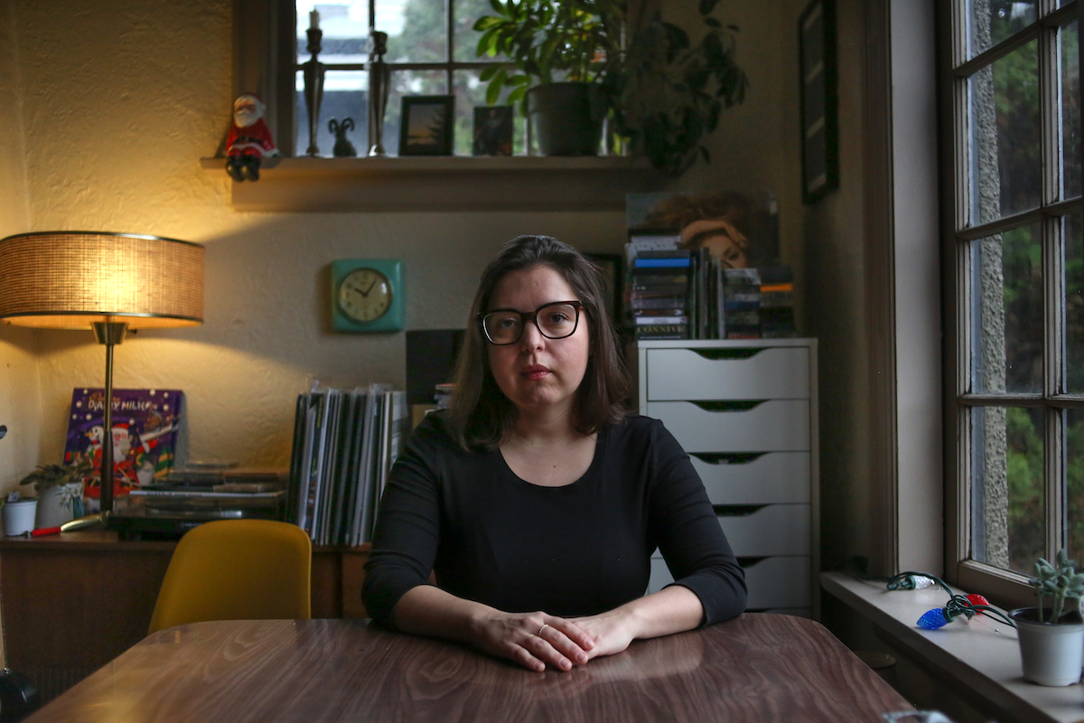 Teresa Alfeld is seated at a Formica table with wood finishings in her apartment beside a window to the right of the frame. She has shoulder-length dark hair and glasses with a thick dark frame, and she is wearing a black long-sleeved top. Beside her is the back of an empty mustard-coloured chair, and to the left of the frame is a mid-century lamp on a tabletop featuring an advent calendar, a record player, vinyl records and a vintage turquoise clock.