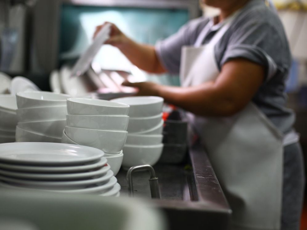 In the foreground, stacks of white dishes in a restaurant kitchen, with a person in soft focus washing dishes in the background.