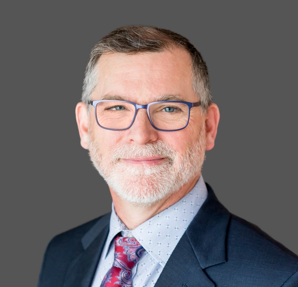 Headshot of a man with light skin, salt-and-pepper hair and a white beard wearing a dark jacket, patterned shirt and paisley tie.