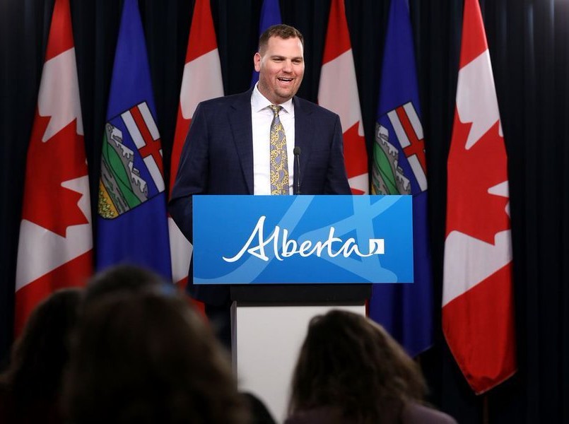 A man with light skin and short brown hair wearing a dark suit speaks at a podium that reads 'Alberta.'