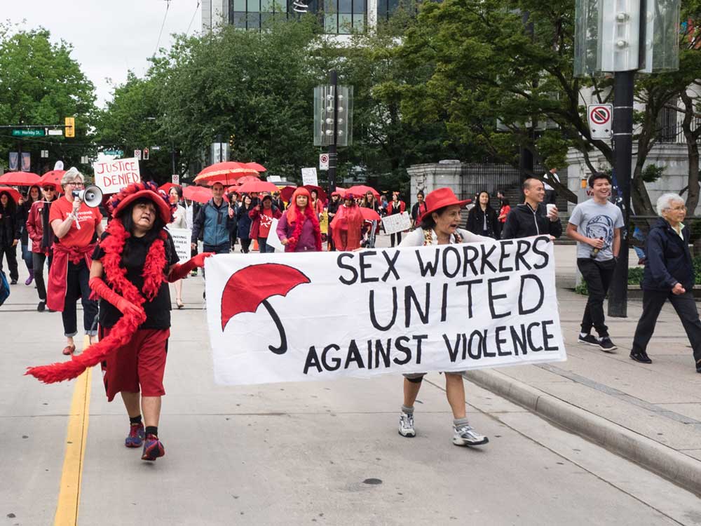 People wearing red and holding red umbrellas march in Vancouver holding a sign that reads 'Sex Workers United Against Violence.'